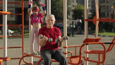 Granddaughter-and-grandfather-doing-fitness-exercises-with-dumbbells.-Senior-man-with-child-kid-girl