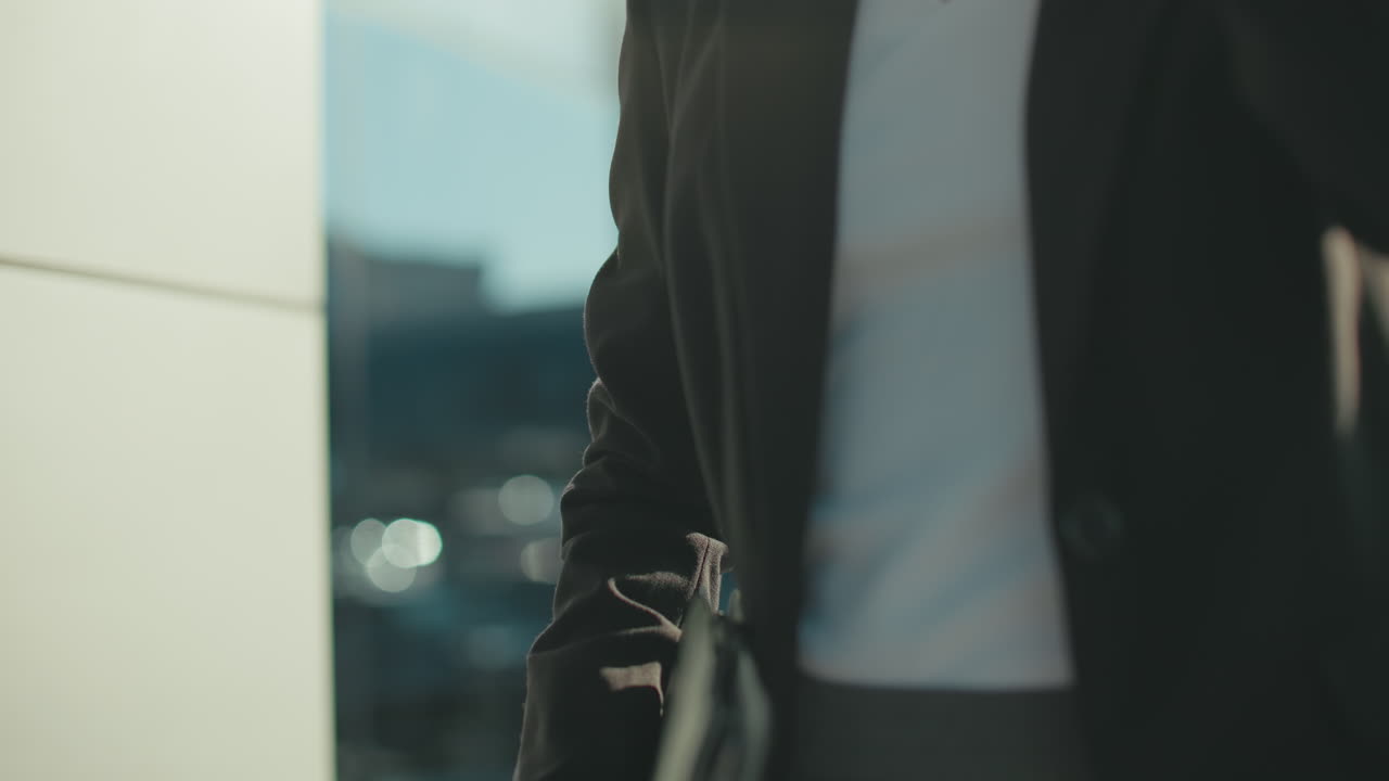 Close up of woman in business attire holding notepad and coffee cup drinks while walking by glass building reflecting blurred cityscape in sunlight with focus on hand gesture and beverage