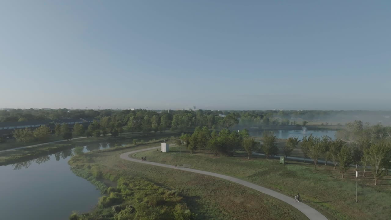 A 4K aerial view of a young boy riding his bike, with a family and other adults walking under clear skies, with ground fog and mist at Exploration Green in Clear Lake, Houston, Texas.