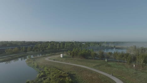 a 4k aerial view of a young boy riding his bike, with a family and other adults walking under clear skies, with ground fog and mist at exploration green in clear lake, houston, texas.