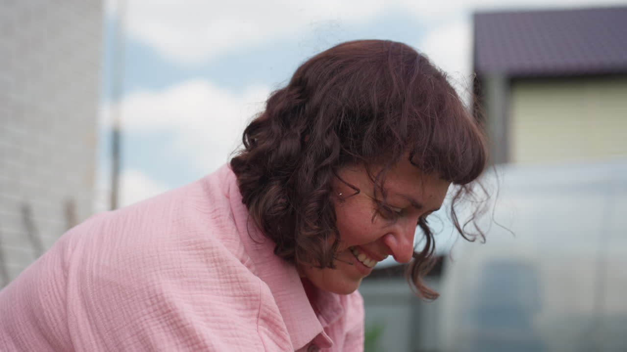 Closeup Woman Smiling In Backyard Leaning Forward, Curly Hair And Pink Shirt, Greenhouse And House In Soft Focus Background, Candid Laughter With Mischievous Expression, Warm Daylight, Authentic