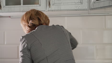 back view of house help wiping white kitchen tiles with green cloth in modern bright home interior maintaining cleanliness hygiene showing daily chores effort