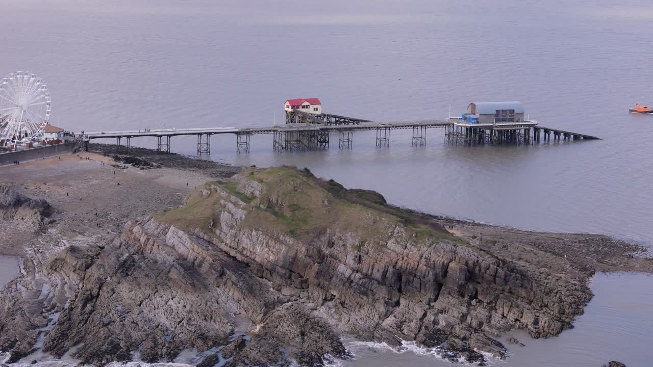 Overhead drone tracking left along Mumbles Pier, to reveal the Big Wheel, rolling waves, and coastal scenery