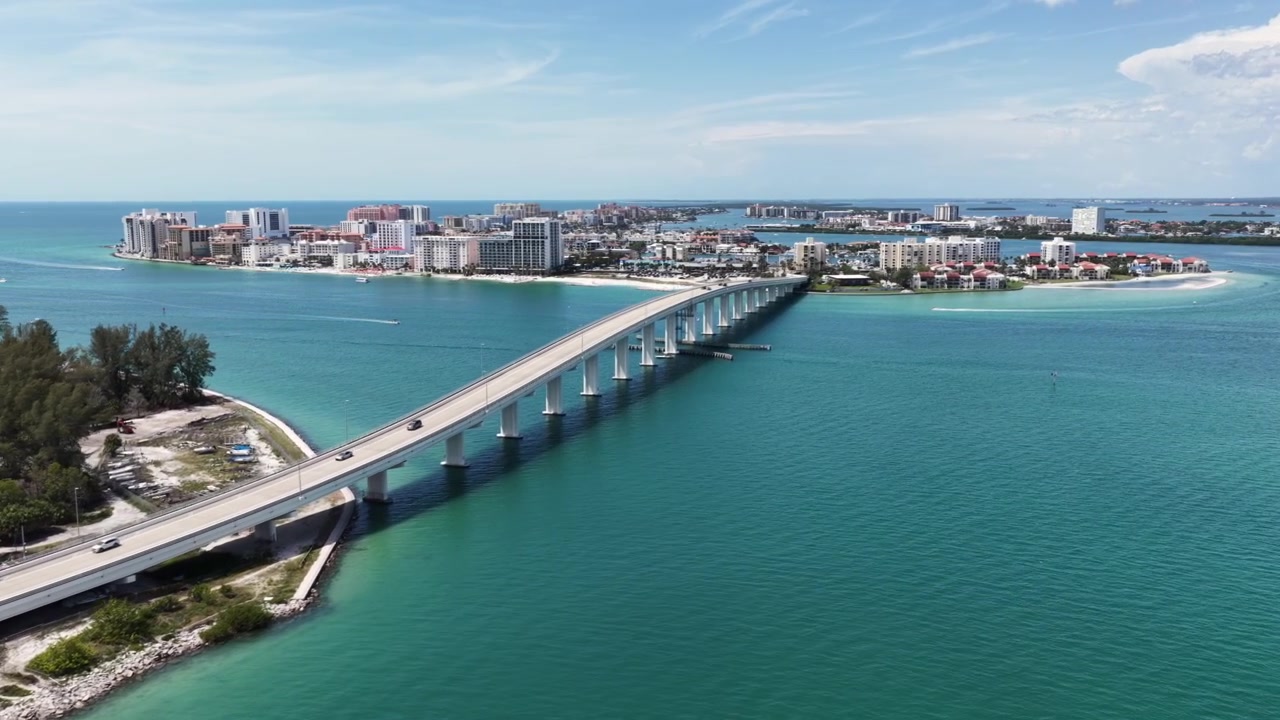 Un ponte rialzato attraversa acque turchesi collegando una spiaggia fiancheggiata da palme allo skyline di Clearwater, con barche e condomini lungo la costa distante. Clearwater Beach, Florida, USA. Ampia ripresa aerea con drone.