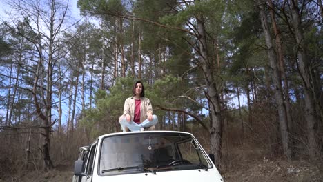 a pretty brunette girl is sitting relaxed on top of the roof of a caravan in the middle of the forest.