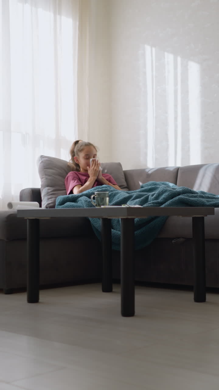 Girl with flu lying on couch wrapped in blanket, holding tissue and looking exhausted, covering herself as she rests in sunlight streaming through window, trying to recover from illness