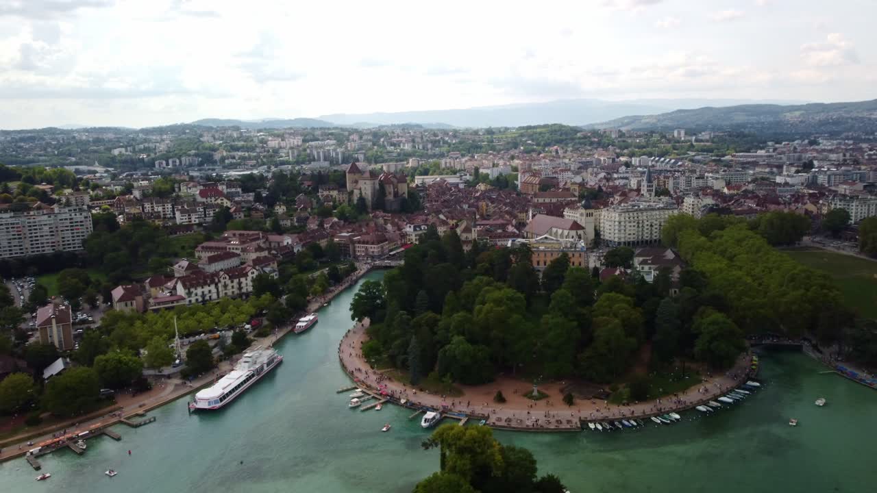Aerial footage over Annecy in Southern France. Footage moves sideways to the right over the ferry port to a park. Views showing the city with buildings stretching into the distance. Boats moored