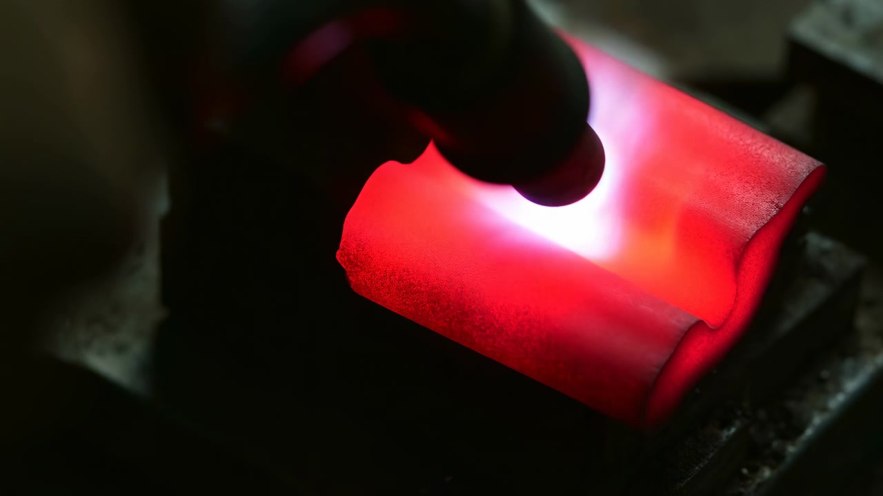 A metal object glows intensely under the heat of a torch in an indoor industrial workshop, creating a warm and industrious atmosphere with vibrant red and orange hues. The close-up, topdown.
