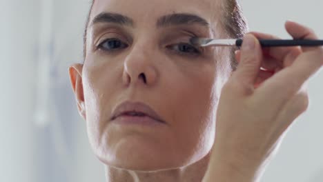 middle-aged woman doing makeup with brush in front of mirror at home