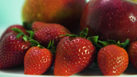 Las-Fresas-Maduras-Frescas,-Grandes-Y-Sabrosas,-Y-Las-Manzanas-Rojas-Cubiertas-Por-Gotitas-De-Agua-Giran-Lentamente-Sobre-Un-Plato-Blanco-Sobre-Fondo-Azul-Claro,-Concepto-De-Comida-Saludable,-Primer-Plano-Extremo,-La-Cámara-Gira-A-La-Izquierda