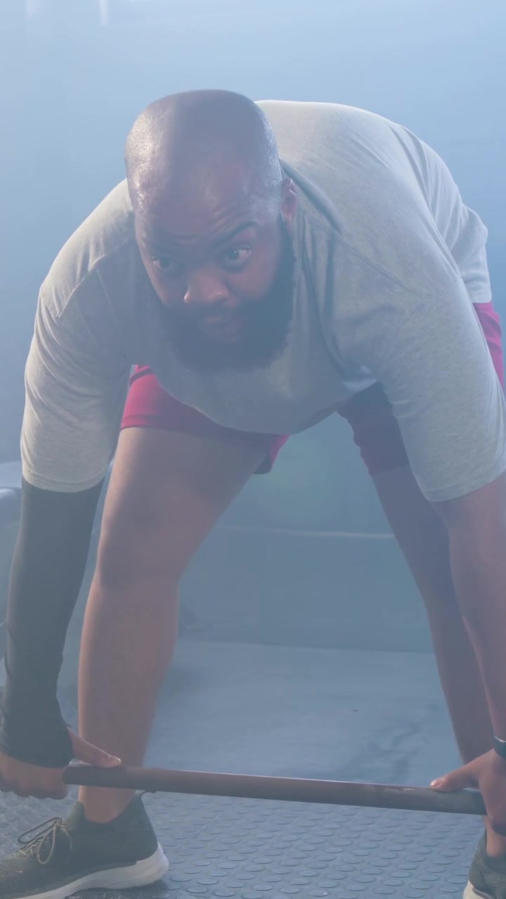 Vídeo vertical: Homem flexionando, com camiseta cinza e shorts vermelhos, contraindo o abdômen, levantando barra de metal e treinando na academia.