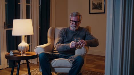 pensive senior man sitting on armchair drinking alcohol from glass and thinking about life in his living room at night, loneliness and solitude concept