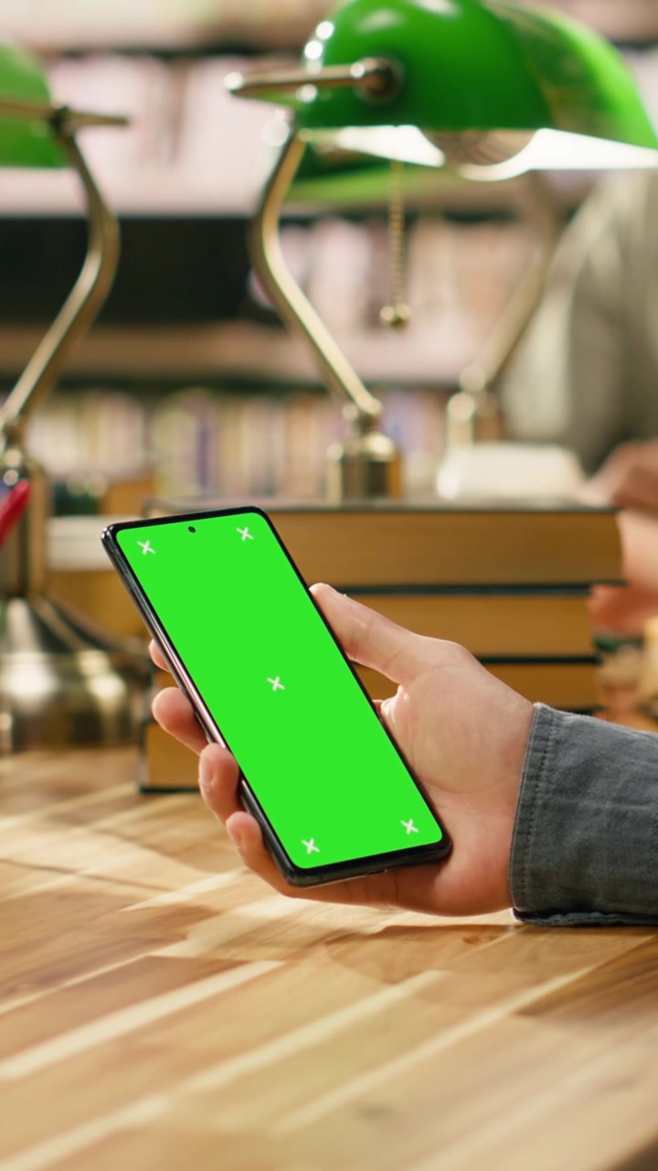 Video verticale: Schermo verde verticale isolato su smartphone tenuto sopra una scrivania di legno della biblioteca