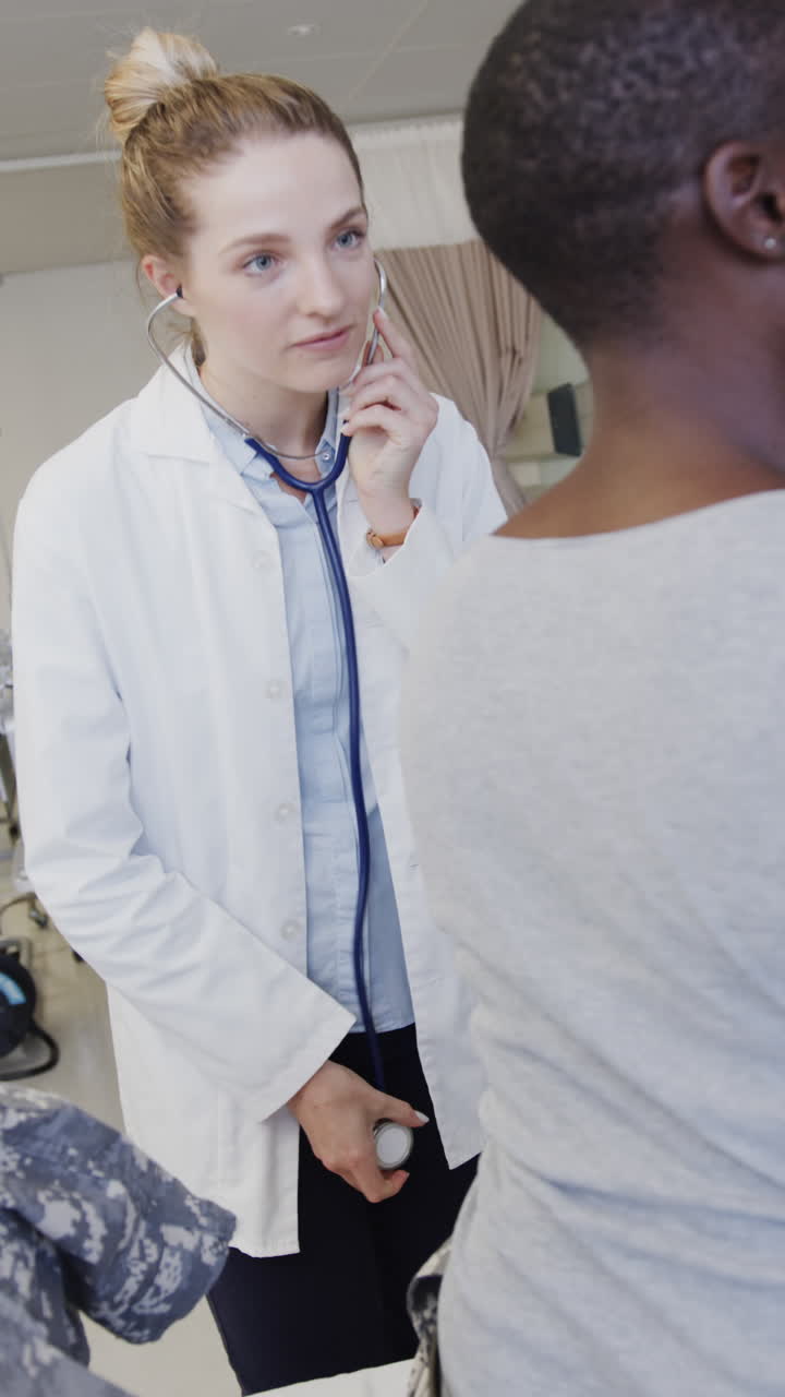 Vertical video of happy diverse female patient and doctor using stethoscope on back, slow motion