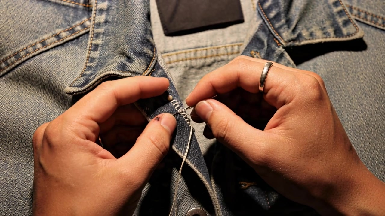 Tangan yang terfokus menjahit plaket denim di meja, menarik jarum dan benang untuk memperbaiki tepi.