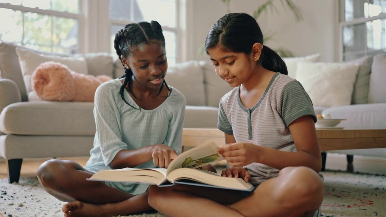 Turning right-side child flipping picture book to read, pointing left child in blue top at home