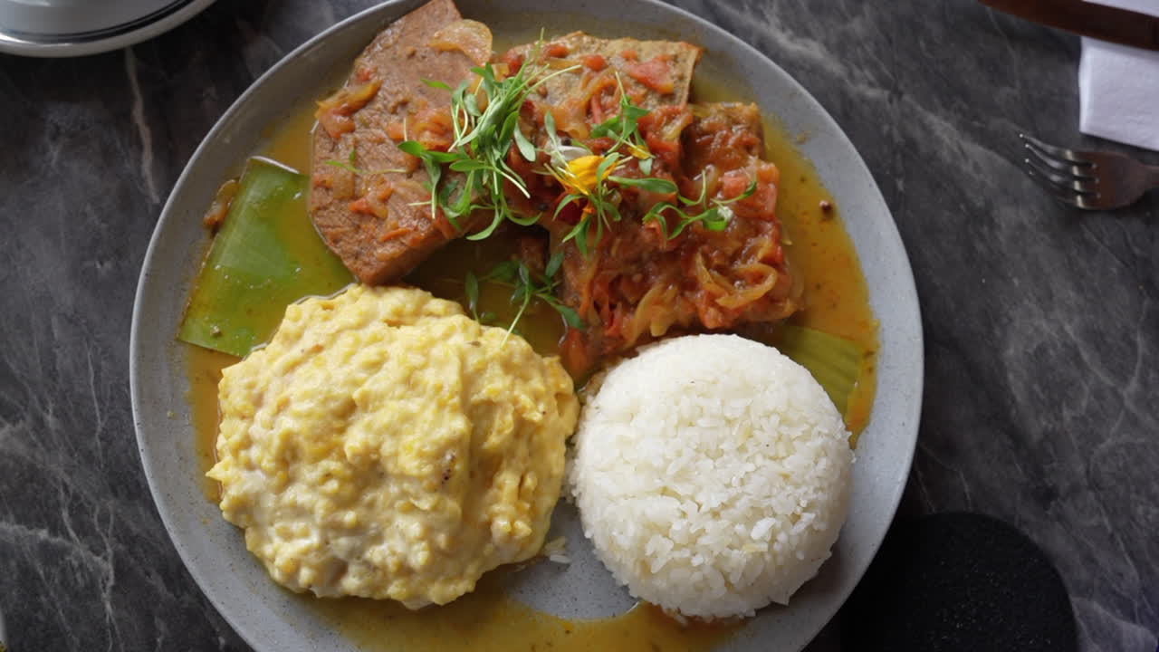 A delicious Colombian feast of sobrebarriga topped with savory sauce, creamy plantain mash, and fluffy rice. Vibrant plating on a textured marble table, showcasing local flavors