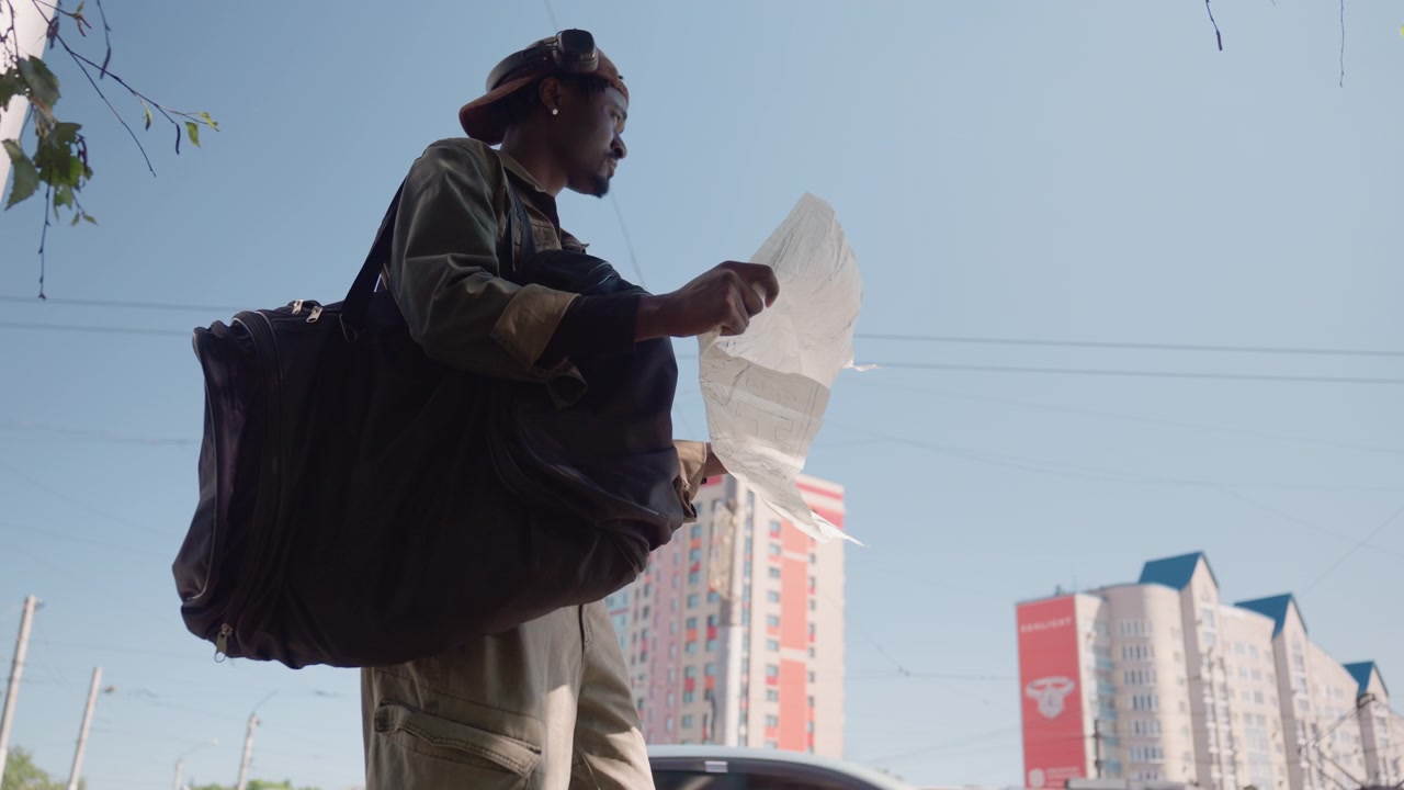 Man wearing cap and carrying large bag examines papers while walking outdoors in urban environment with tall buildings under clear blue sky. Man holds documents close to his body suggesting