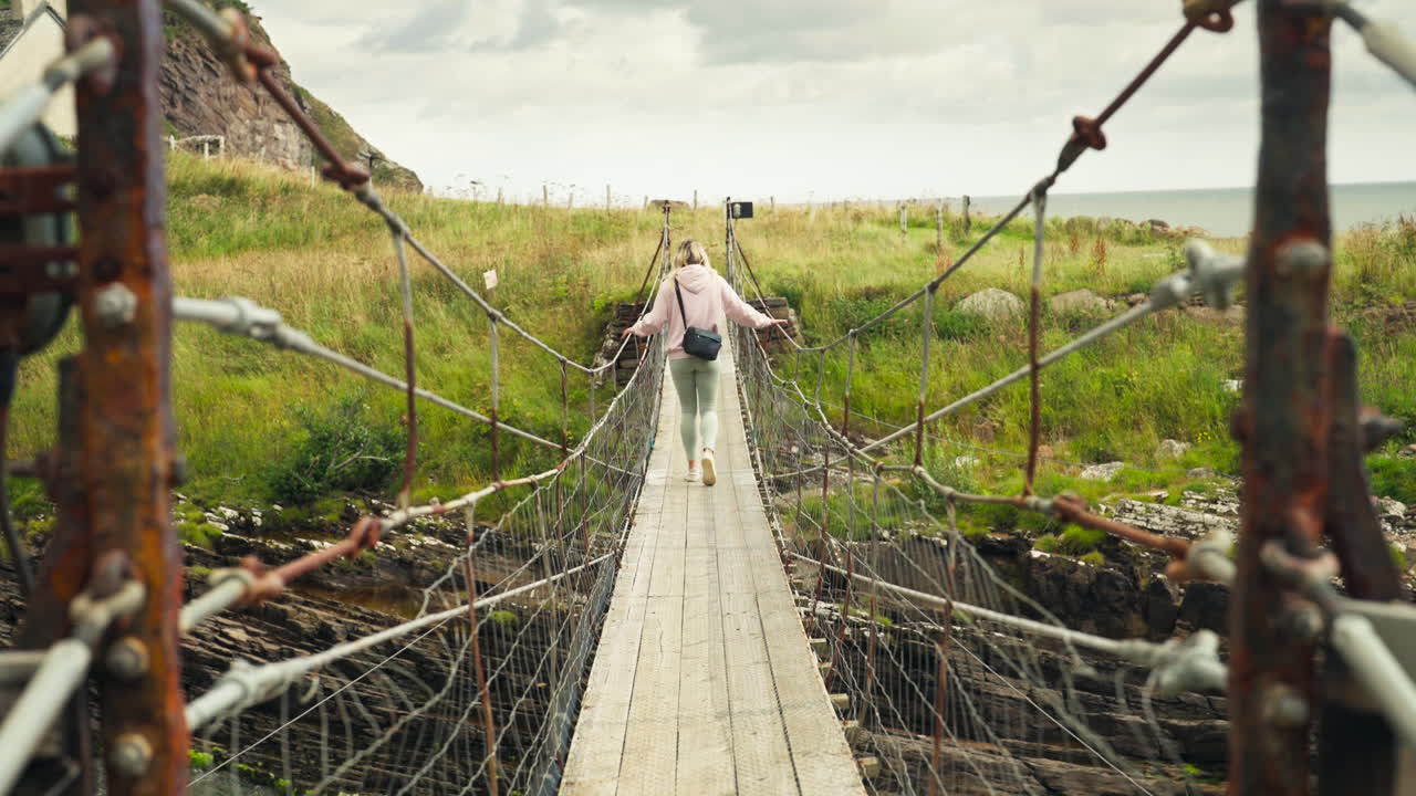 Handheld shot of blond caucasian woman crossing a suspension bridge in the day in Scotland, United Kingdom