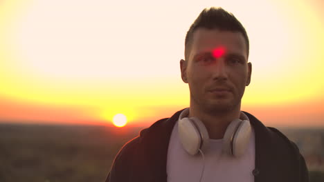 Smiling-and-laughing-handsome-young-man-listening-music-from-his-smartphone-in-wireless-headphones.-In-slow-motion-a-man-stands-on-the-roof-at-sunset-and-looks-at-the-beautiful-view-from-the-height-of-the-city