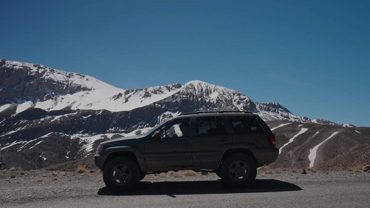 Fas'taki Atlas Dağları'nda karla kaplı zirvelerin altında çakıllı yolda park etmiş koyu renkli SUV.