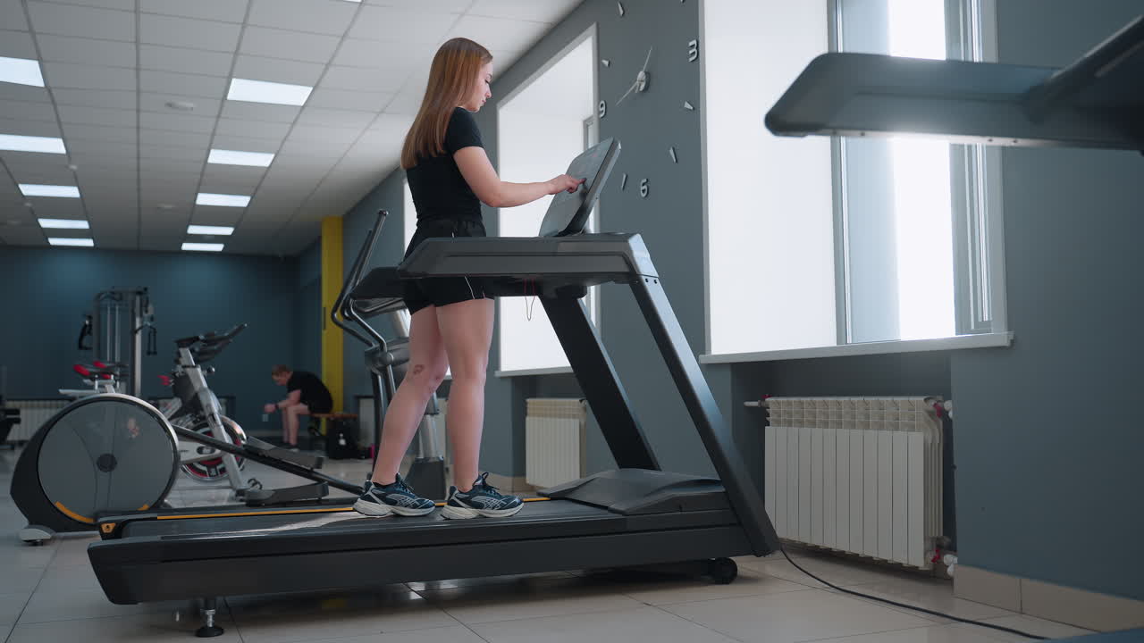 woman in black gym outfit walks on treadmill adjusting digital console under sunlight filtering through large window in modern gym studio with exercise bikes and elliptical trainer visible behind