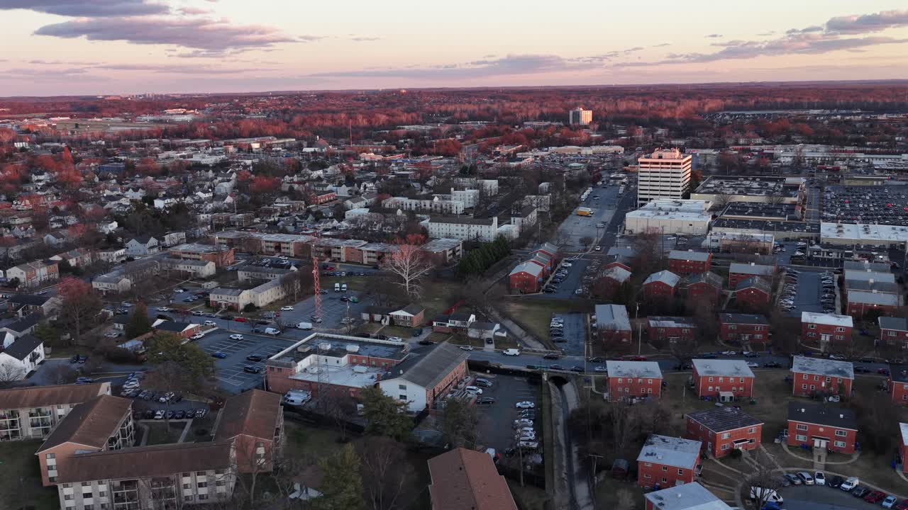 Aerial sunset flyover of Laurel, Maryland, showing residential neighborhoods, apartment complexes and commercial buildings glowing in warm winter light under calm evening sky. Wide shot