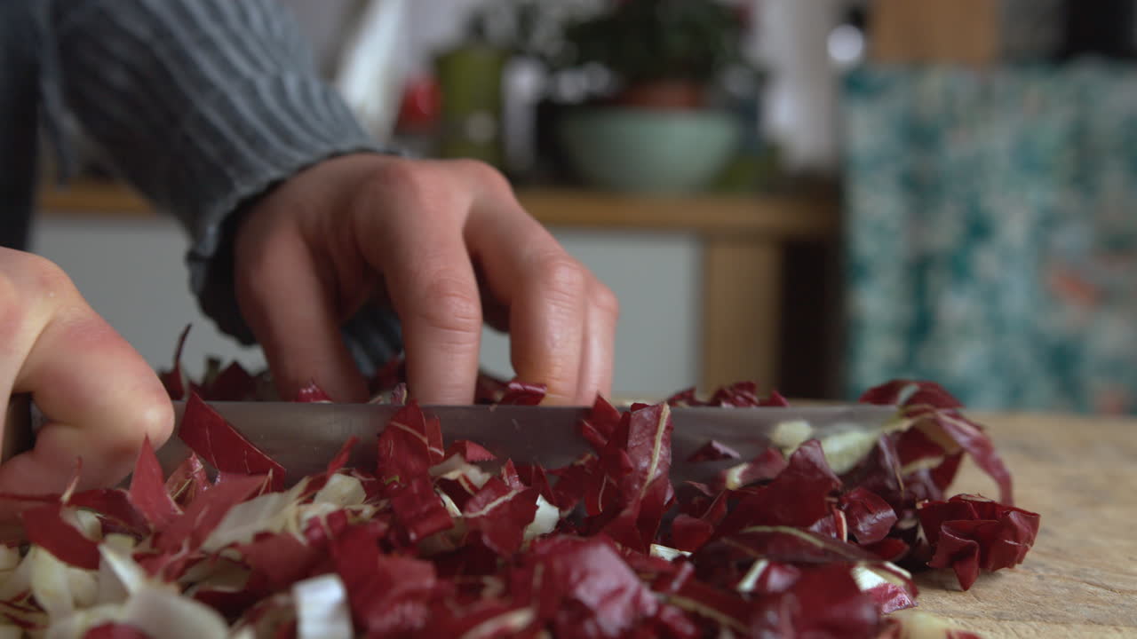 крупным планом наклонная женская рука аккуратно нарезает острым ножом итальянский цикорий на деревянной доске на кухне