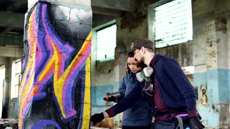 professional graffiti artist is teaching pretty girl to paint with aerosol spray paint standing together inside adandoned warehouse near old column and talking.