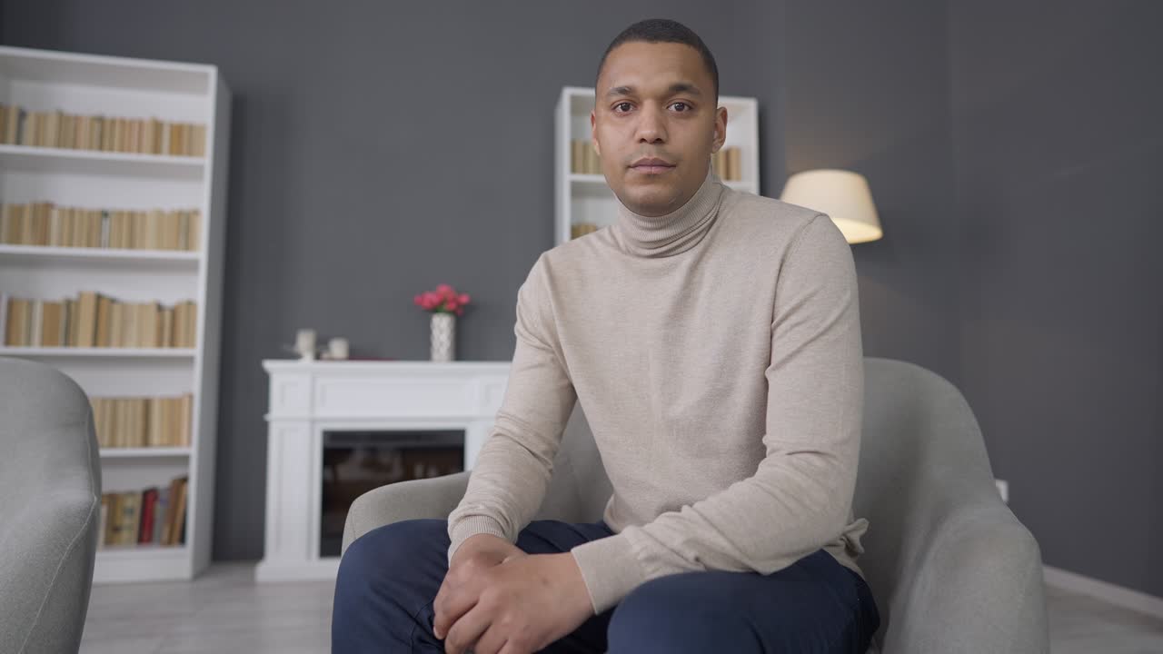 Confident young African American man sitting on comfortable armchair at home looking at camera. Portrait of successful handsome smart guy posing indoors. Lifestyle and confidence concept.