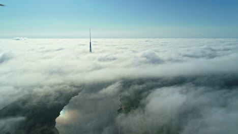 Aerial-view-of-a-foggy-clouded-landscape-with-the-top-of-a-radio-or-TV-antenna-in-blue-sky