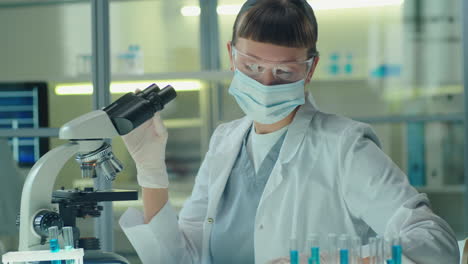 female scientist in mask and glasses working with microscope in lab