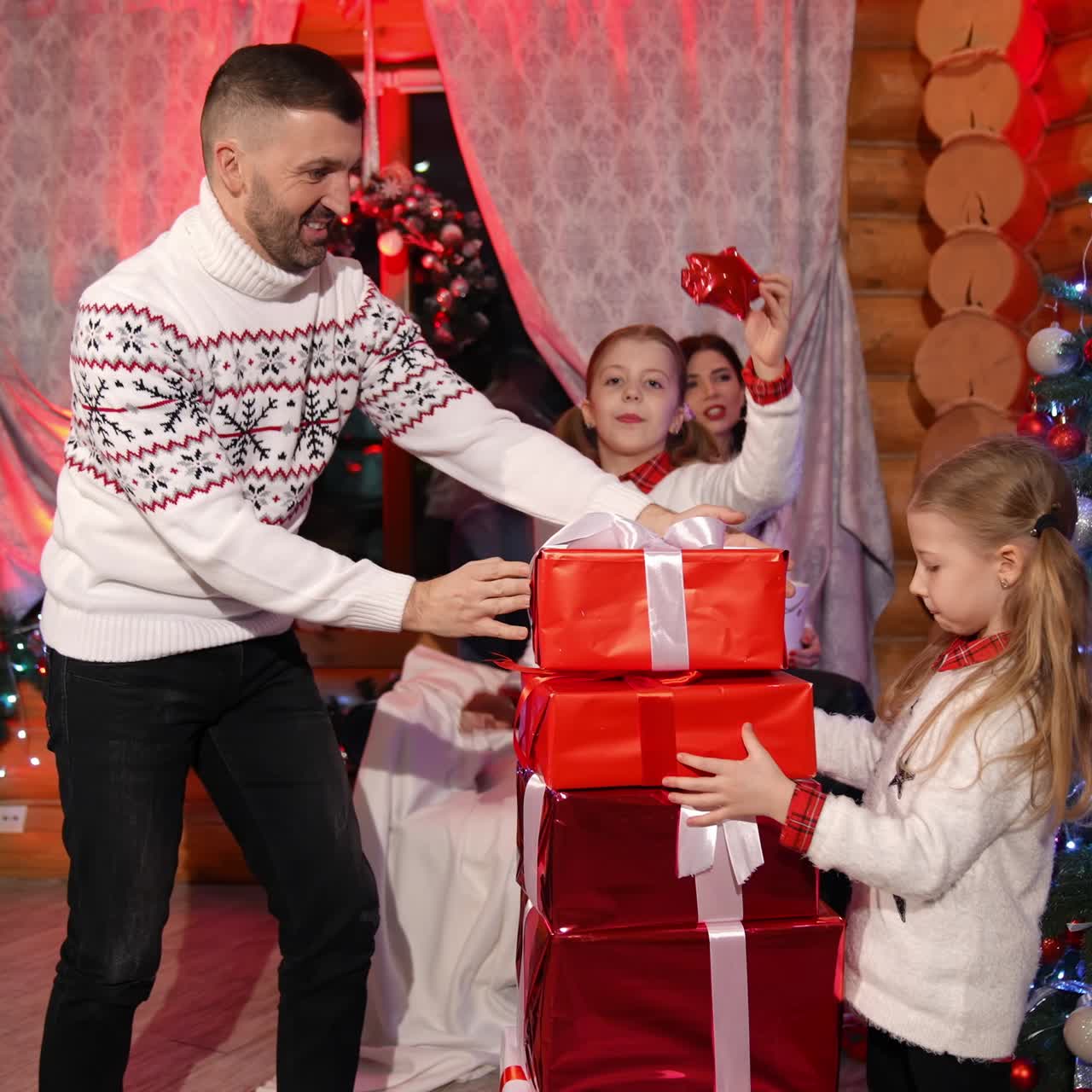Father and two daughters are placing presents in a tall pile. Smiling mother is watching her family at the background. Beautiful family in decorated room
