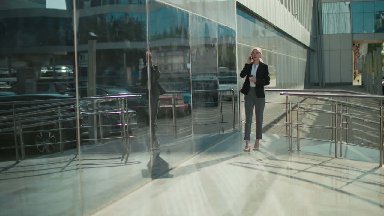 Elegant business woman walking confidently with coffee in hand while on phone call, passing modern glass building with reflections of parked cars and urban surroundings