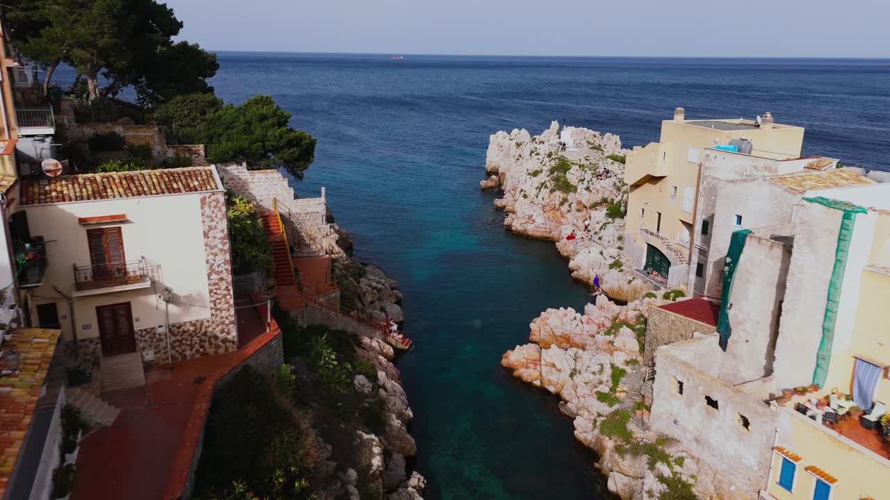 Drone moves slowly forward over colorful rooftops and descends toward a narrow rocky sea inlet in Porticello, Sicily, with clear water and traditional coastal houses visible