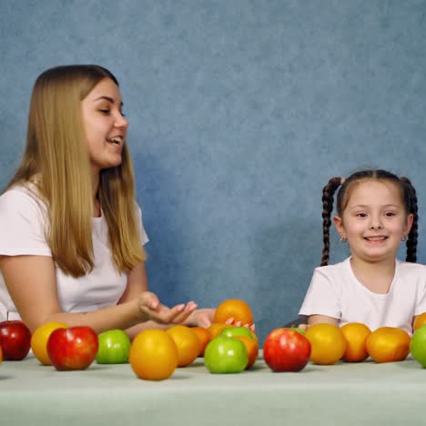 happy mother and daughter play with fruit. beautiful woman and little girl throw up oranges on blue background. fresh organic fruit on the table.