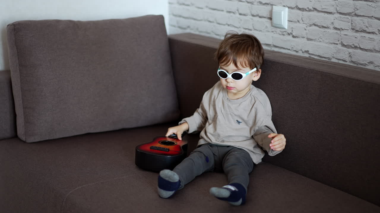 Small Caucasian boy in sunglasses sits on the sofa playing with a little guitar. Future rockstar portrait.