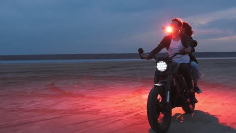 young couple riding on vintage motorcycle with red burning signal fire after sunset on beach, slow motion