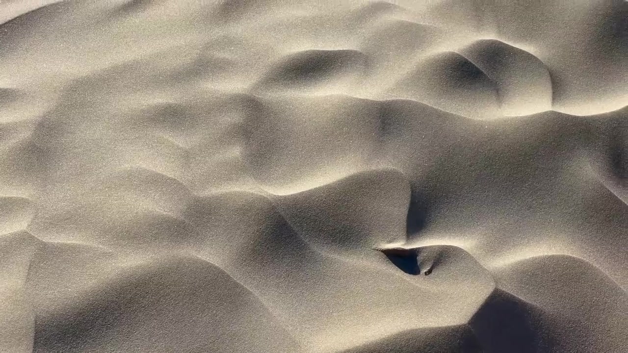 Lateral pan across textured sand dunes with wind-swept patterns and rocks.