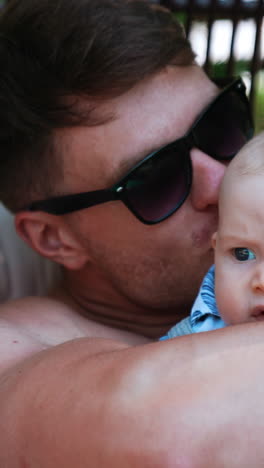 father embracing a cute baby boy in his arms. dad in sunglasses kisses his lovely infant son sitting outdoors. close up. vertical video