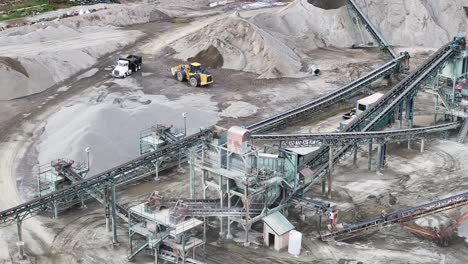 a large gravel manufacturing plant with large piles of rock and a yellow bulldozer tractor dumping a large heap of gravel into a dump truck aerial orbit