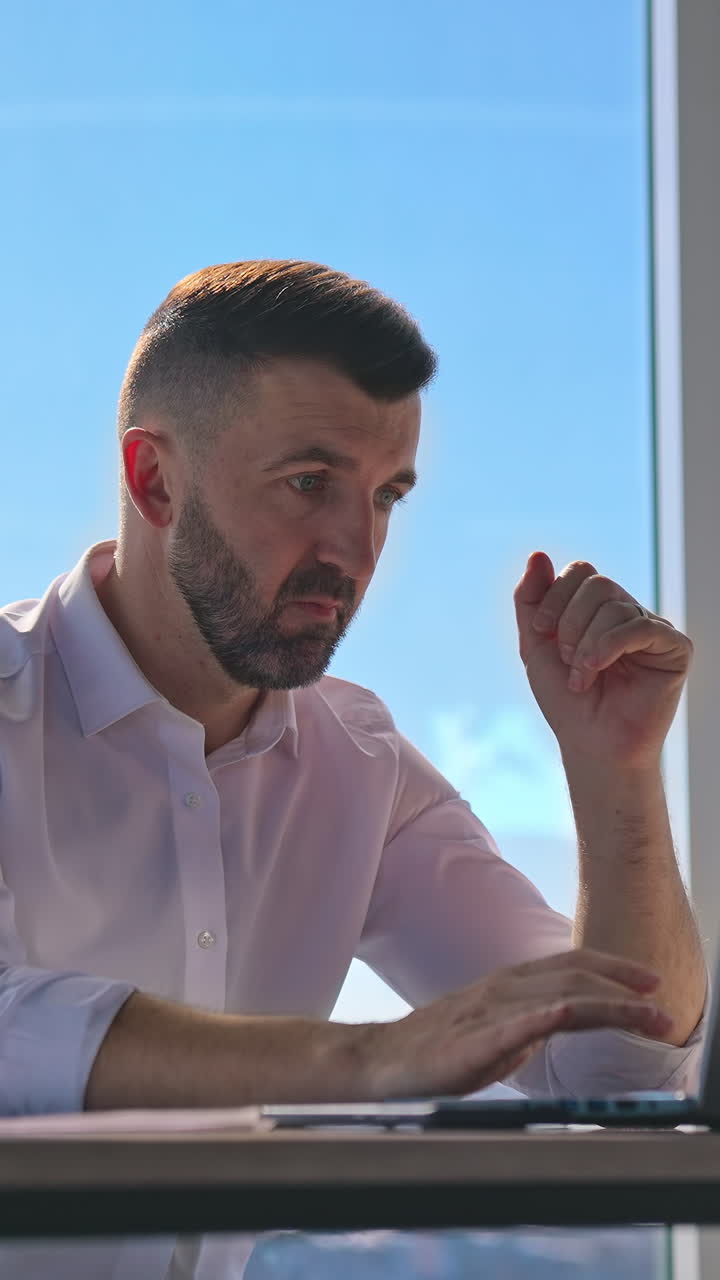 Focused businessman working on laptop computer in office with large windows. Bearded male worker using laptop computer at workplace. Handsome man looking at laptop screen in office. Vertical video