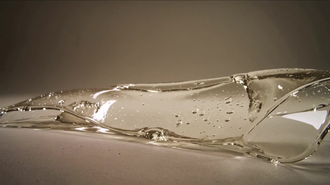 Nastro di gel trasparente che si forma da una cresta, si muove a destra e si posa sul tavolo, con bolle e riflessi, ripresa ravvicinata fissa.