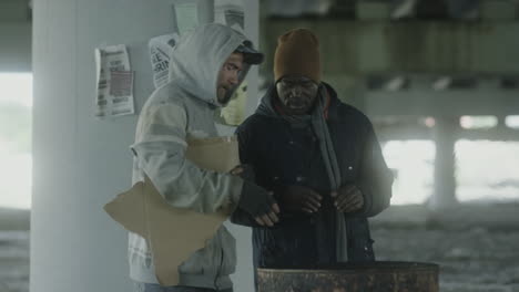 two homeless men huddle around a barrel fire