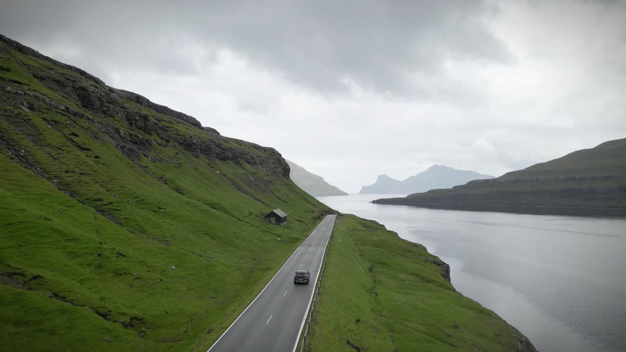 파로 제도 (faroe islands) 에 있는 북대서양의 광활한 넓이와 울창한 푸른 언덕으로 둘러싸인 휘어지는 해안 도로를 따라 운전하는 자동차의 숨막히는 공중 촬영.