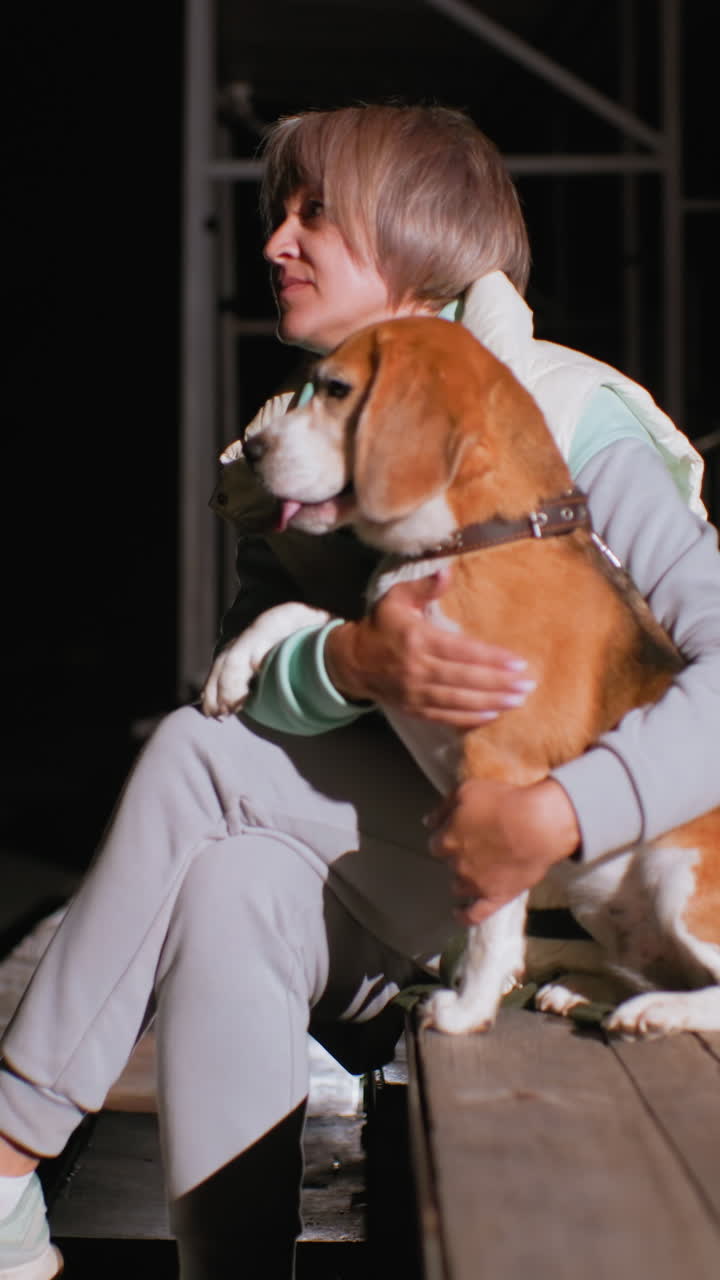 woman white hugging beagle on bench, nighttime urban setting under scaffold, mature female owner cradles panting dog with gentle hands, soft spotlight highlights warm coat and collar, casual tracksuit