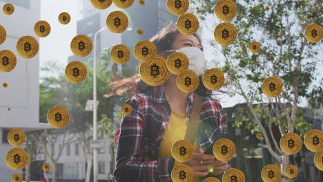 animación de múltiples símbolos de bitcoin sobre mujer asiática que lleva una máscara facial usando un teléfono inteligente
