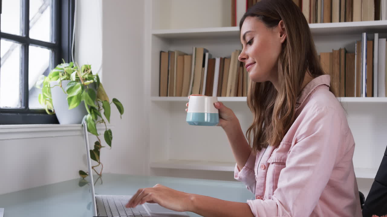 Woman working on laptop at home, holding coffee mug, surrounded by books