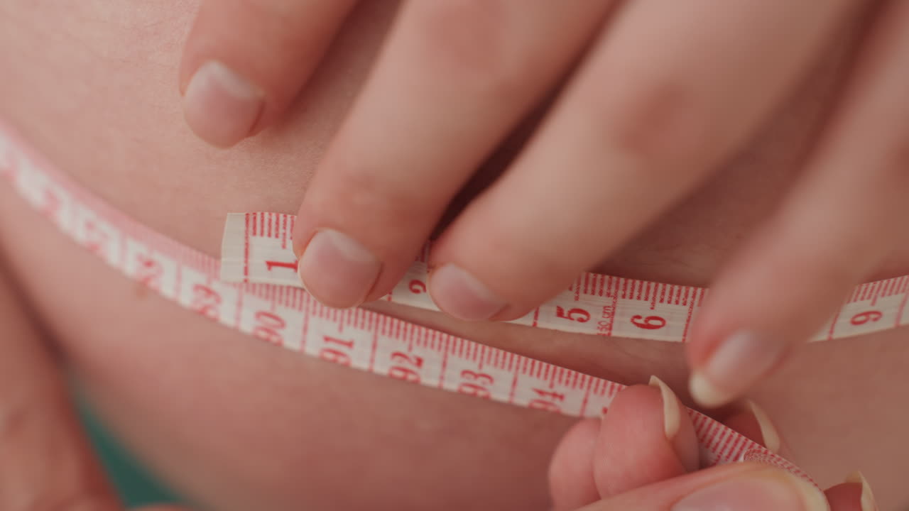 Primo piano estremo della pancia con i numeri del metro allineati, dita che regolano il punto zero, segni dei centimetri visibili, texture della pelle morbida, misurazione precisa della maternità e documentazione delle tappe.