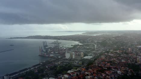 Left-trucking-aerial-drone-ultra-wide-shot-of-The-Port-of-Salvador-located-in-All-Saints-Bay-in-Salvador,-Bahia,-Brazil-on-a-cold-rainy-fall-morning-with-poor-neighborhoods-below
